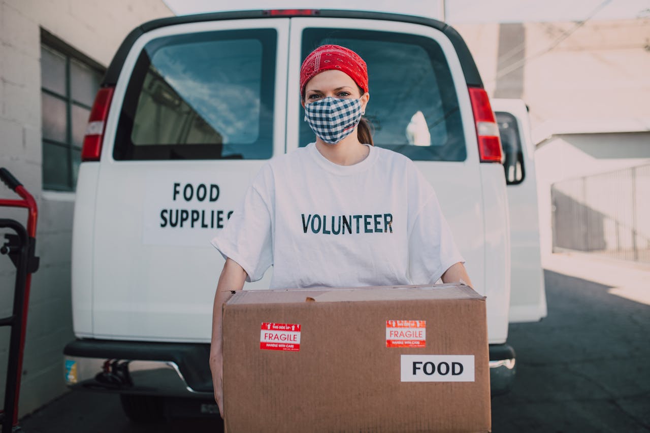 contact-img Volunteer wearing a mask delivering food supplies during the pandemic, holding a carton box labeled 'FOOD'.